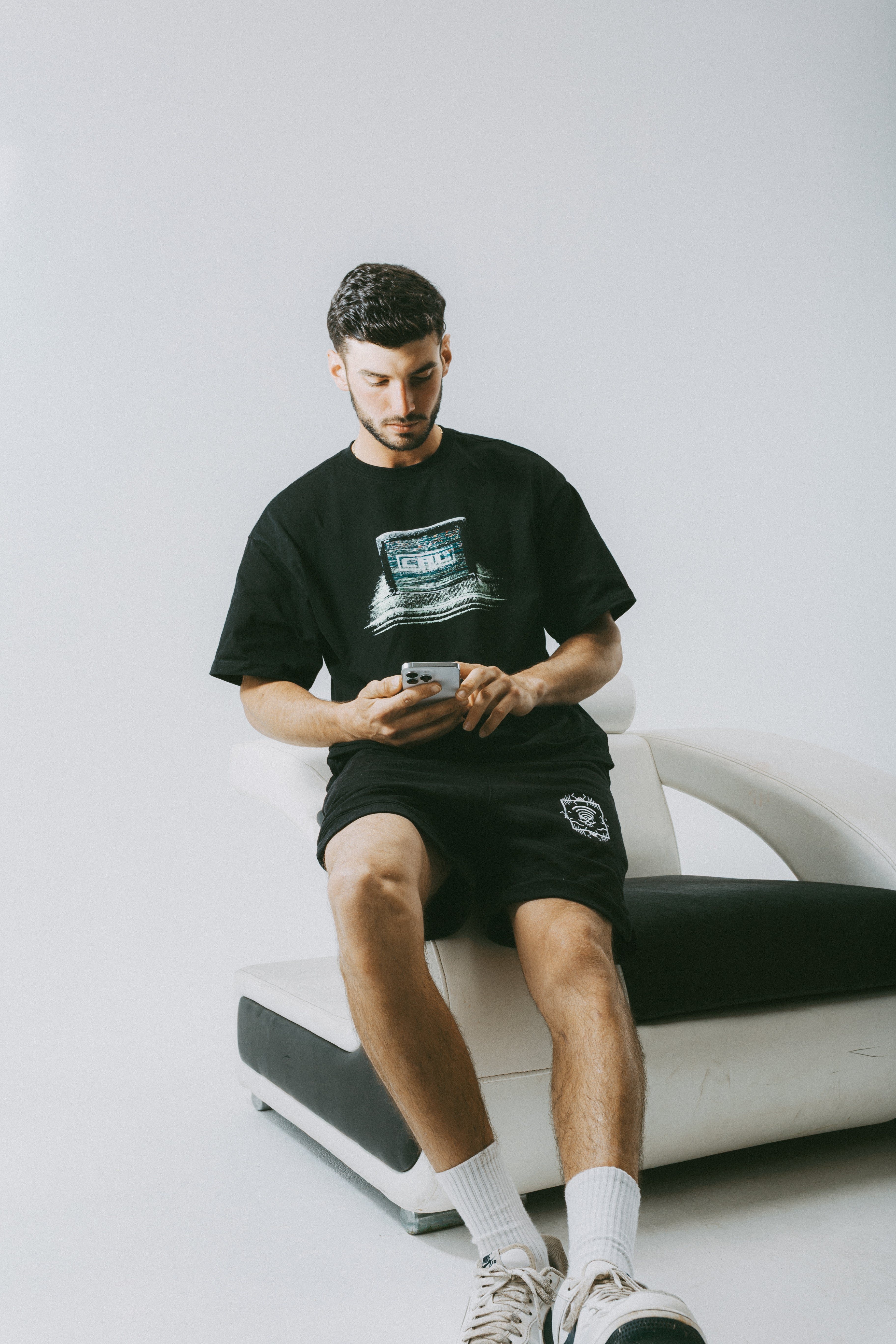 Young man wearing a graphic streetwear tee and shorts, sitting on a stylish chair.